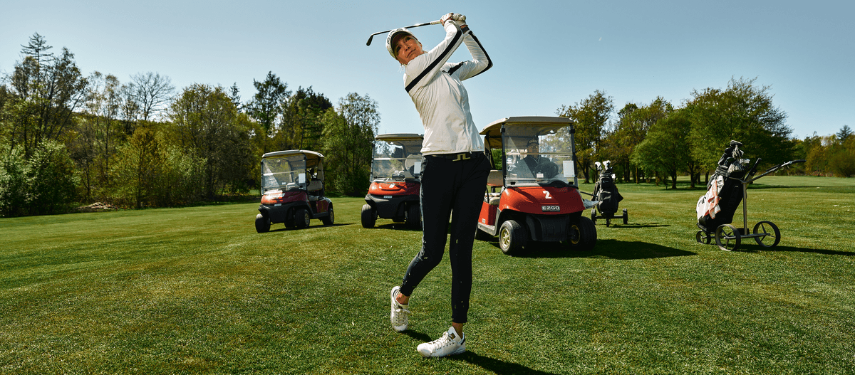 Golferin beim Abschlag auf gepflegtem Grün mit Golfwagen im Hintergrund