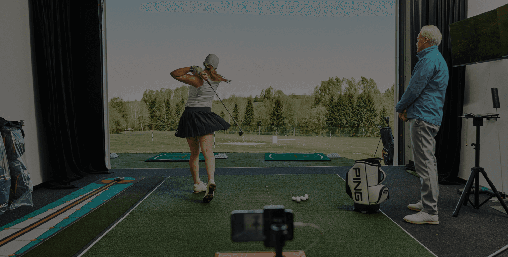 Golferin beim Abschlag in moderner Indoor-Trainingshalle, Coach Martyn Robinson beobachtet.