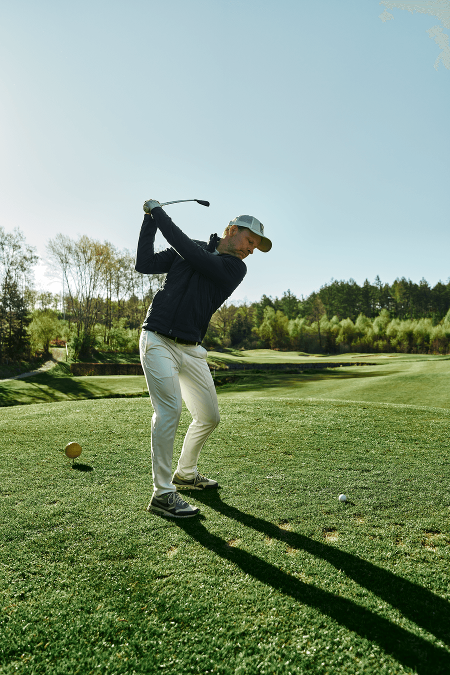 Golfer beim Abschlag auf grünem Fairway vor sonniger Waldkulisse.