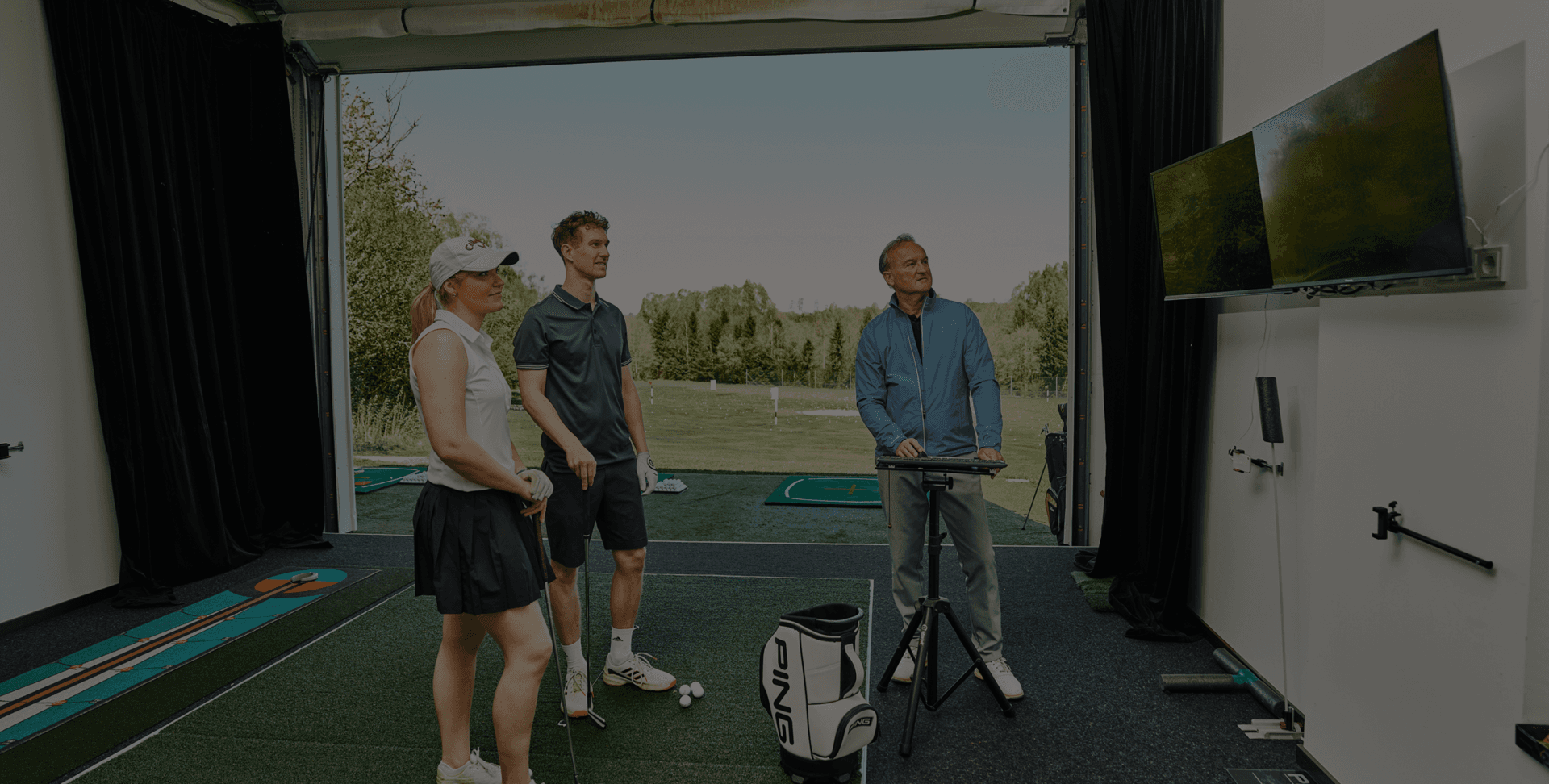 Golfschüler in Indoor-Anlage mit Trainer und Analyse-Bildschirmen vor malerischem Hintergrund