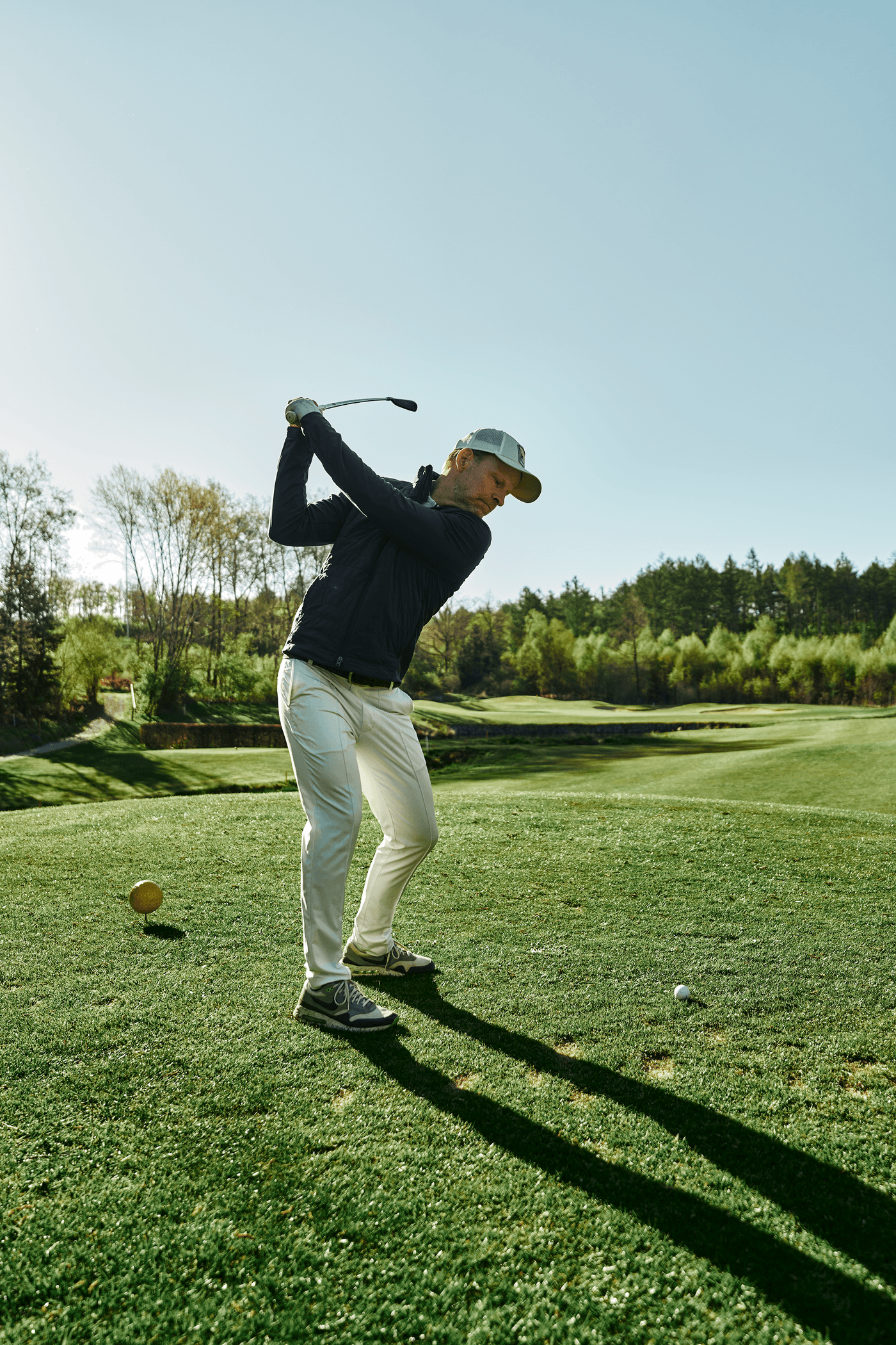 Golfer beim Abschlag auf grünem Fairway vor sonniger Waldkulisse.